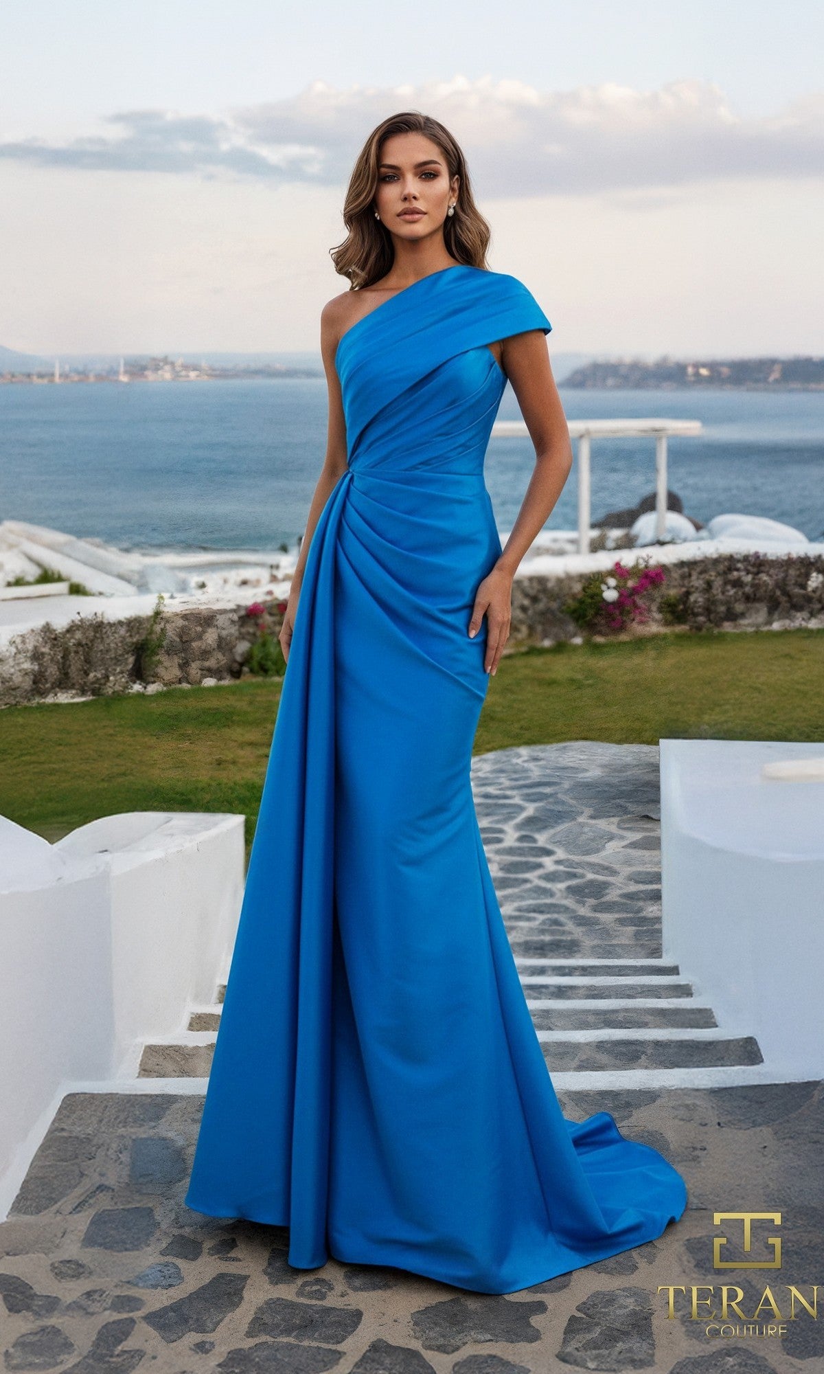A woman stands outdoors on a stone walkway, wearing the Terani 261M6411 long formal dress in Peacock. The elegant one-shoulder gown with draped detailing is set against a seascape and striking white architecture.
